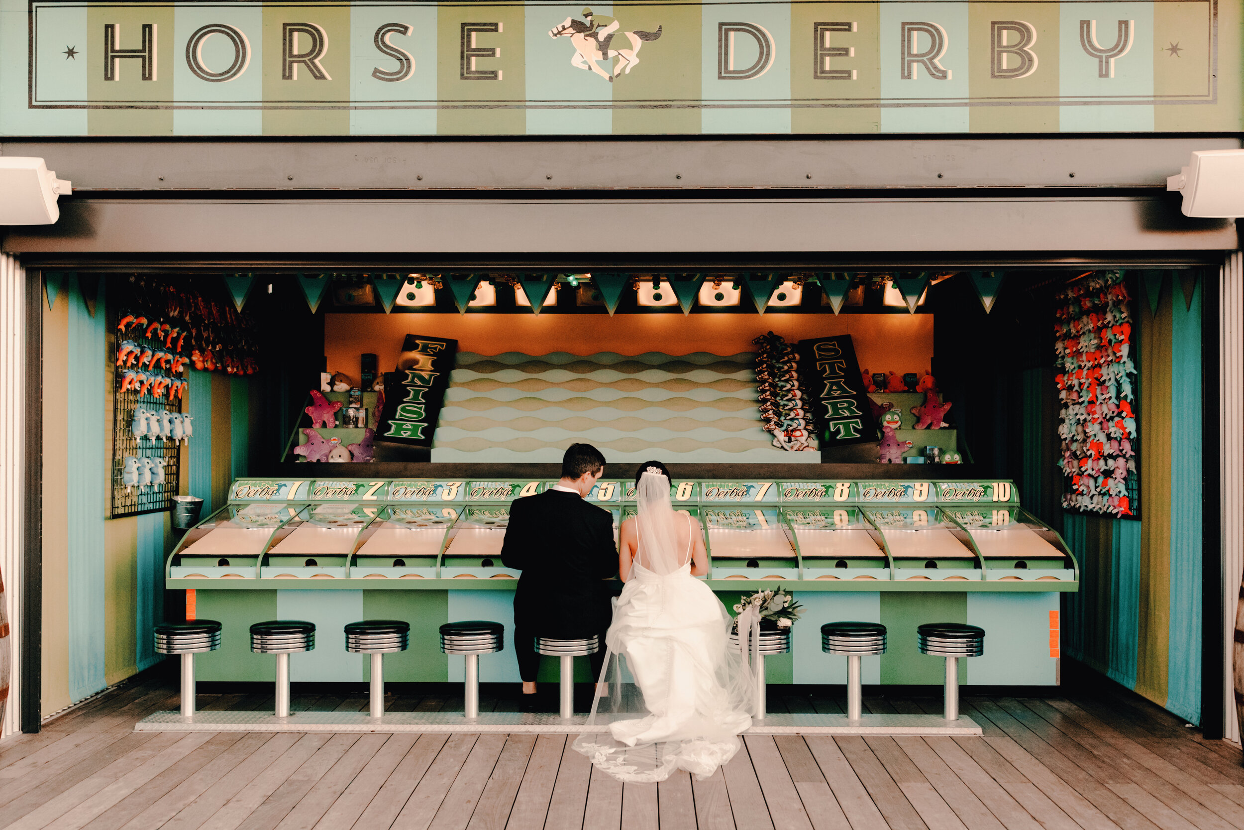 Bride and groom standing in front of a carnival game booth at the Horse Derby fair, with the bride in a white wedding gown and bride in a black suit, facing the game.