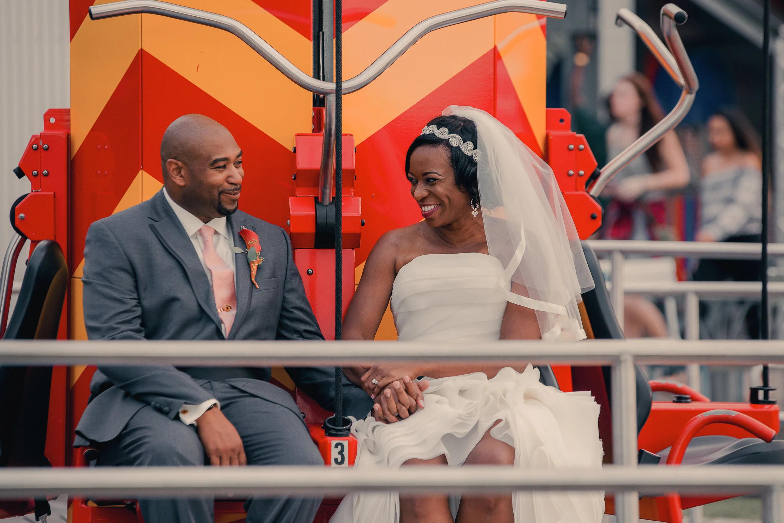A bride and groom sitting on a carnival ride, holding hands and smiling at each other. The bride is wearing a white wedding dress and veil, and the groom is in a gray suit with a pink tie and boutonniere.