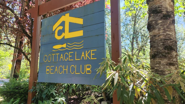 Blue wooden Cottage Lake Beach Club sign with yellow letters among trees in Woodinville