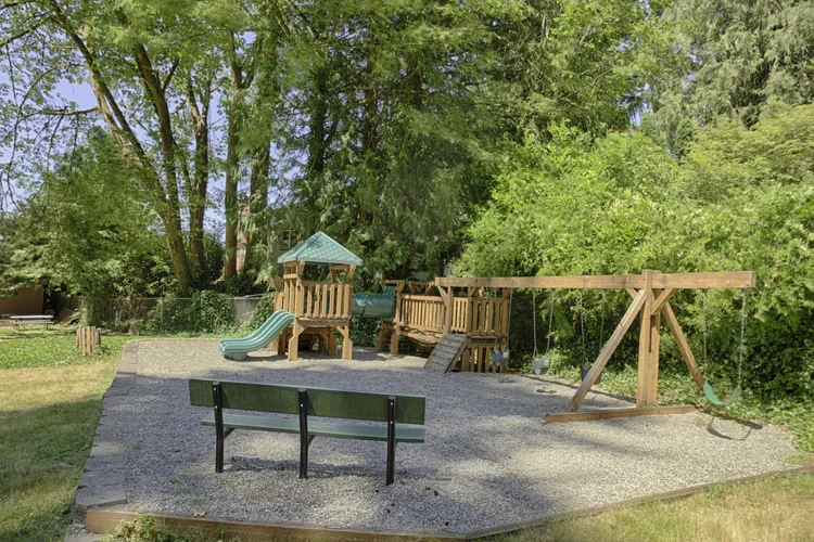 Gravel playground with wooden fort, green slide, swings and a bench at the beach club