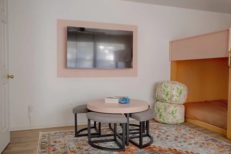 Bunk room lounge with wall TV in a pink frame, round table with stools and floor poufs