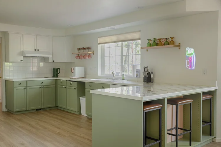 Sage green kitchen with white counters, bar stools, open shelving and a pink neon drink sign