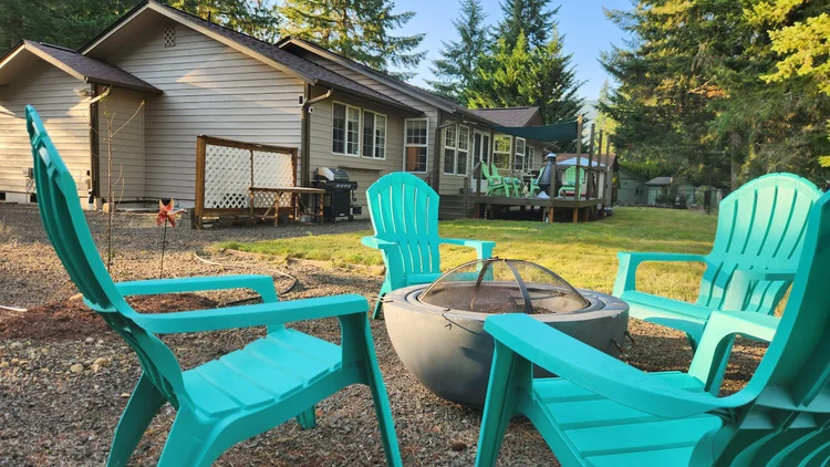 Teal Adirondack chairs around a fire pit on gravel, with the house and deck behind
