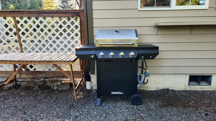 Black three-burner gas grill on gravel beside the house and a wood potting bench