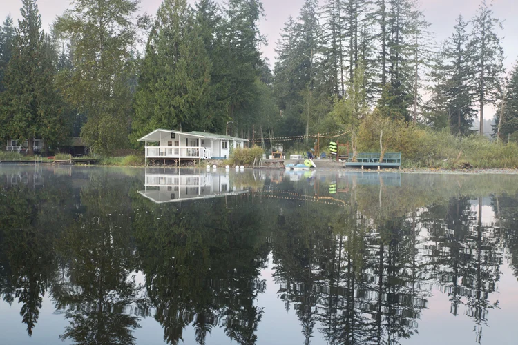 Lakeside Wonderland cottage on a calm Graham lake, framed by tall evergreens in morning mist