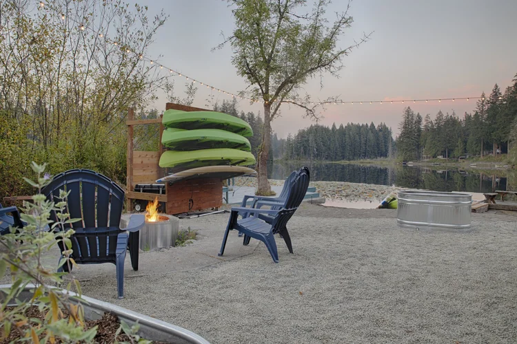 Gravel beach with blue Adirondack chairs around a lit fire pit and a stacked kayak rack