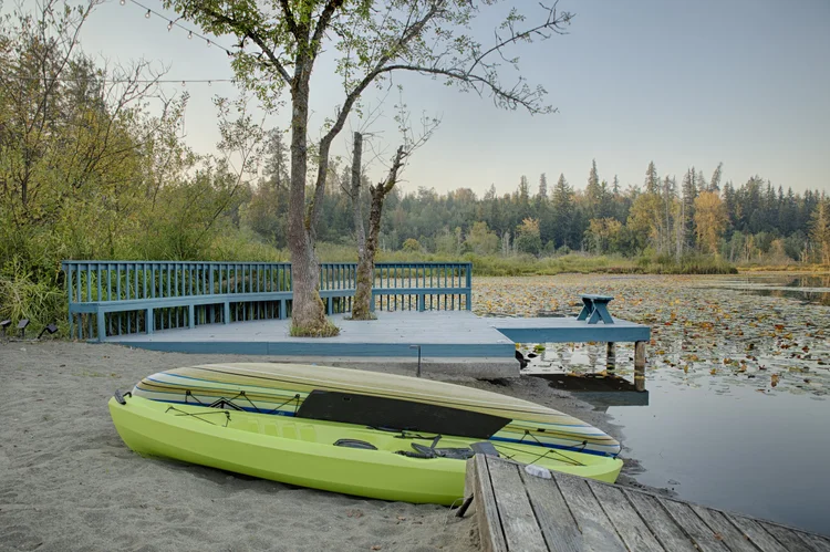 Green kayak and paddleboard on the sand beside a blue-railed dock over a lily pad lake