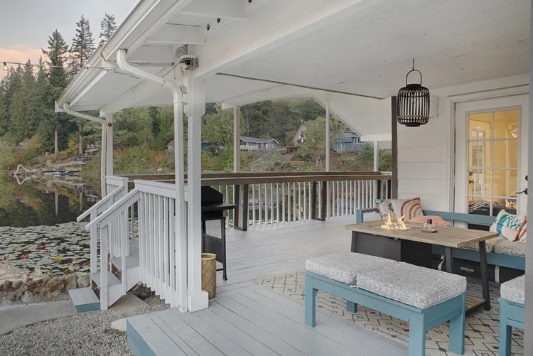Covered white deck with fire table, blue benches and grill beside the lake at dusk
