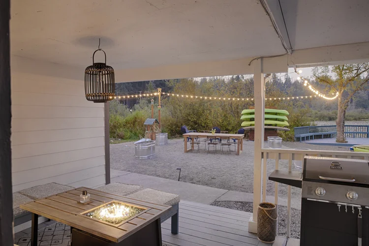 Covered patio fire table and grill facing a gravel yard with string lights and picnic table