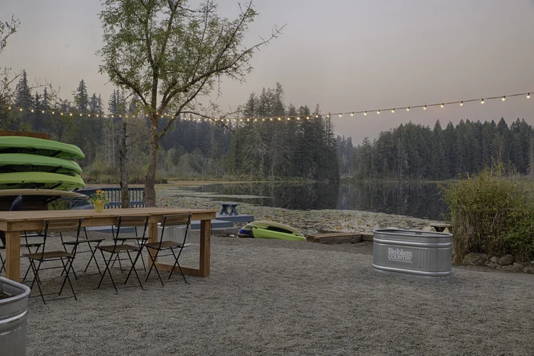 Gravel yard with wood dining table, bistro chairs, string lights and the lake at dusk