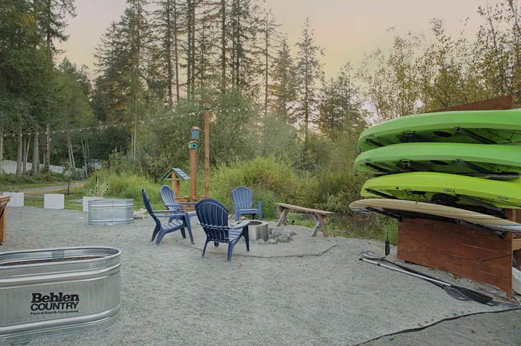 Gravel yard with blue Adirondack chairs around a fire pit beside a green kayak rack