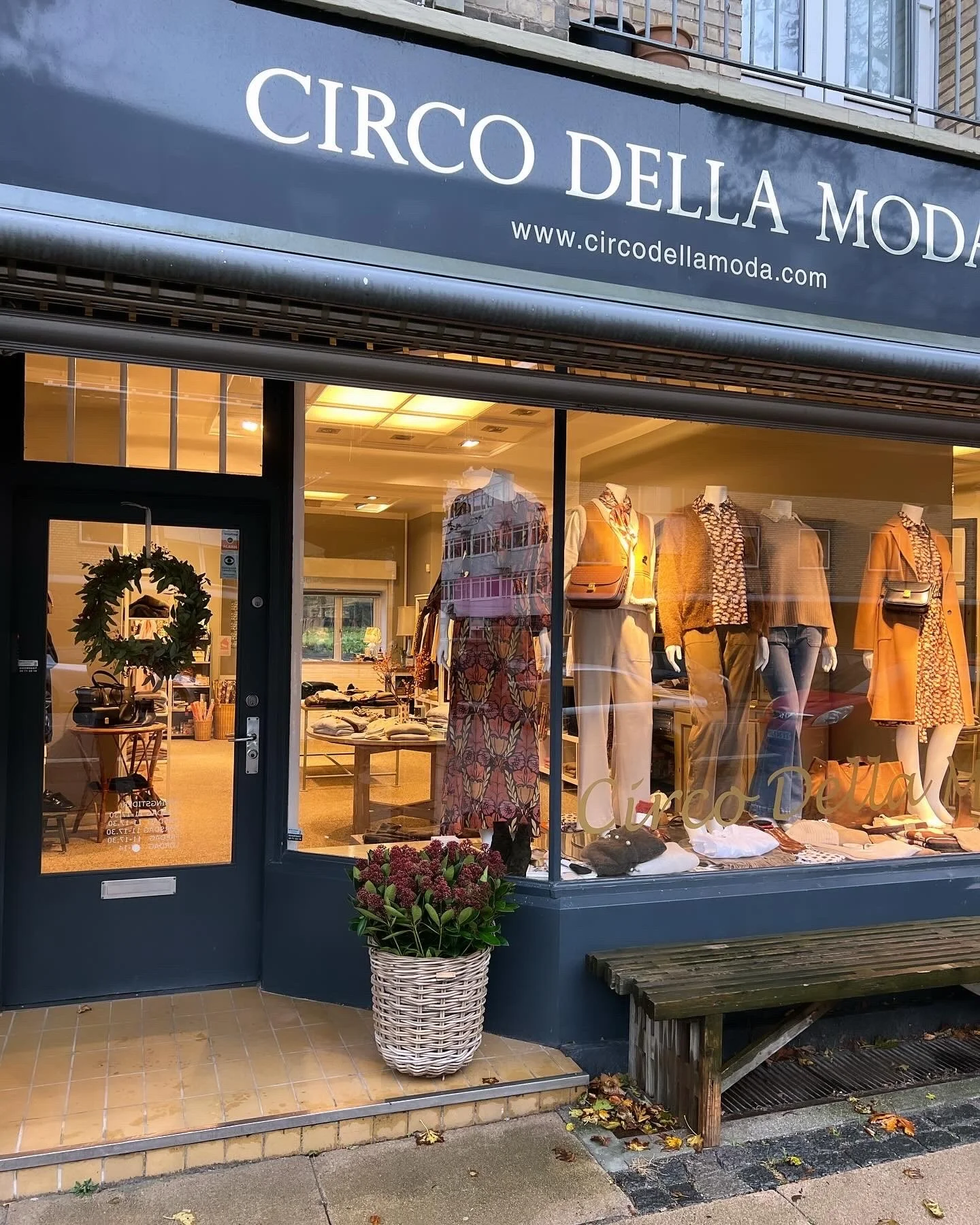 Udendørs udsigt til en tøjbutik med vinduesudstilling af mannequener med tøj, en dør med en julekrans, en stor blomst i en kurv, en bænk foran butikken og nogle efterårsløv på jorden.