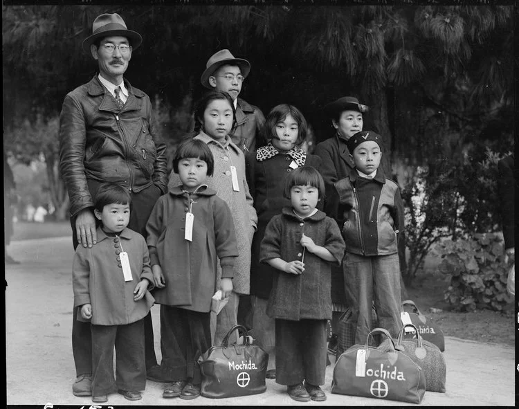 May 8, 1942. Hayward, California. Members of the Mochida family awaiting evacuation bus. Identification tags are used to aid in keeping the family unit intact during all phases of evacuation. Mochida operated a nursery and five greenhouses on a two-acre site in Eden Township. He raised snapdragons and sweet peas. Evacuees of Japanese ancestry will be house in War Relocation Authority centers for the duration. National Archives.