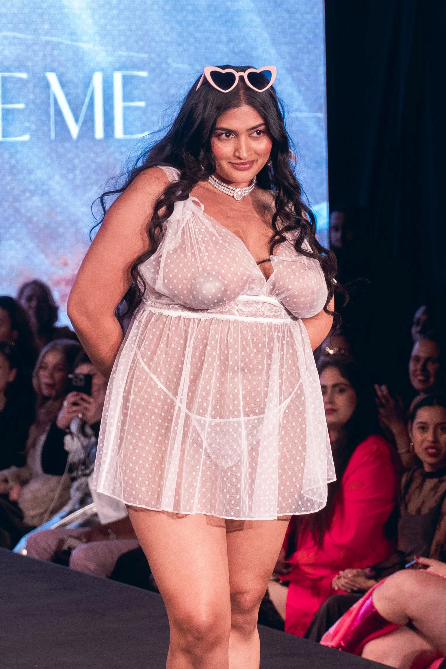 Model in a white sheer mini dress and sunglasses poses on the Adore Me runway at New York Fashion Week 2024
