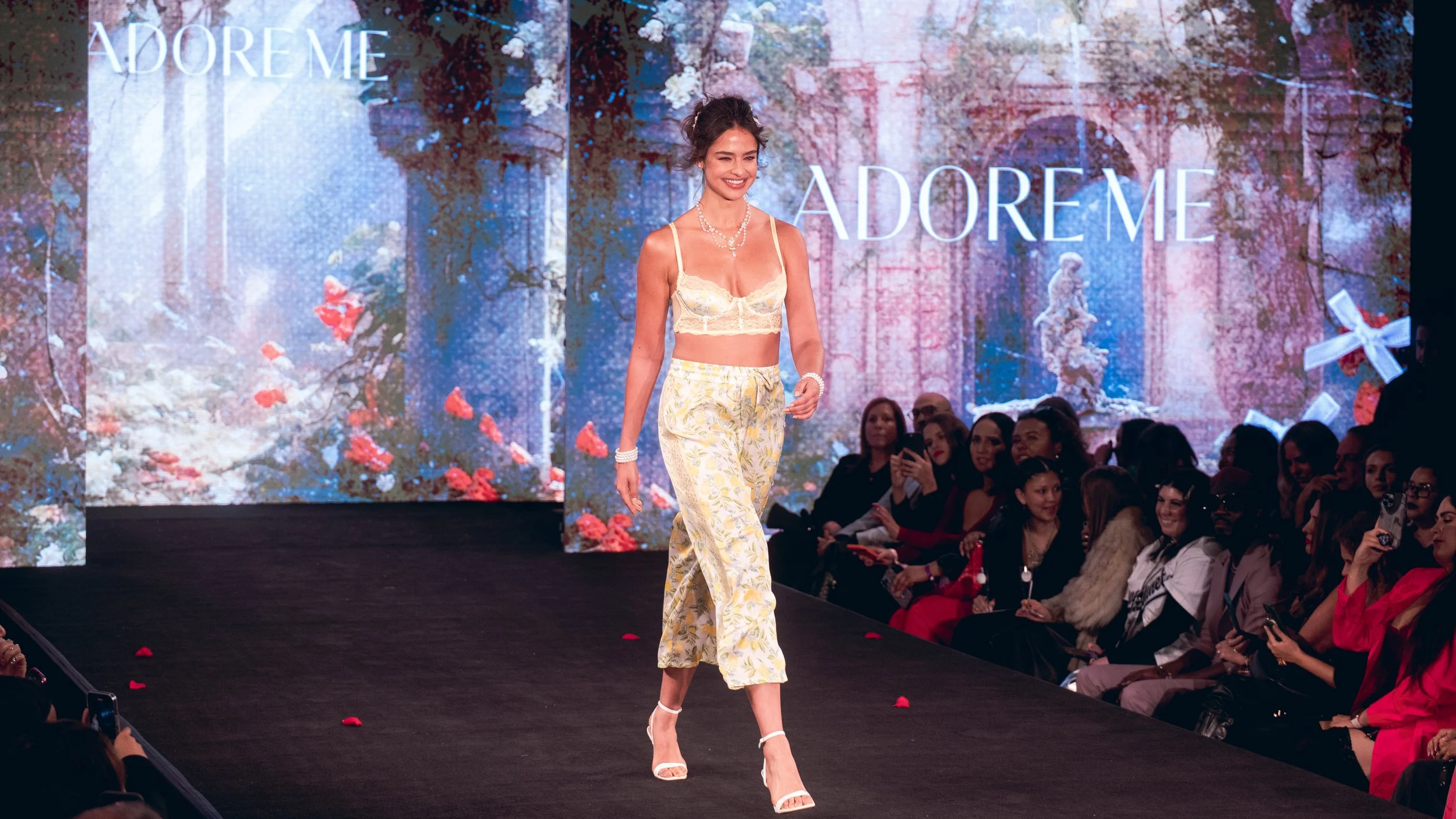 Smiling model in a cream lace top and floral pants walks the Adore Me runway at New York Fashion Week 2024