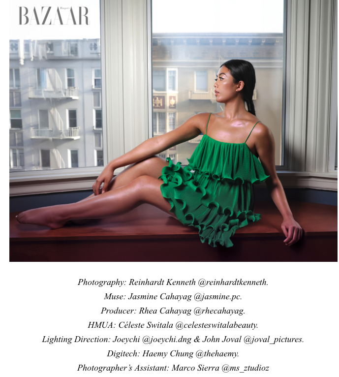 A woman in a green ruffled dress sitting on a windowsill with city buildings in the background, looking to the side.