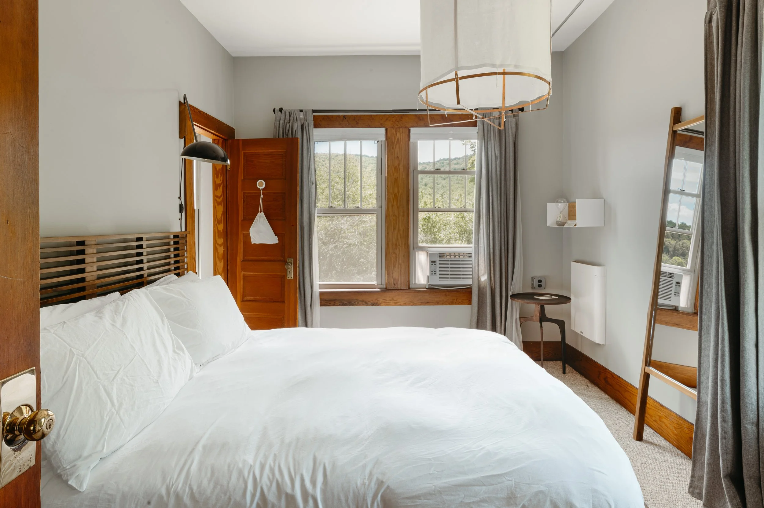 A cozy bedroom with a large bed covered in white bedding, wooden window frames, and a view of green hills outside. The room features a small side table, a mirror, and window air conditioning units, with curtains on the windows.