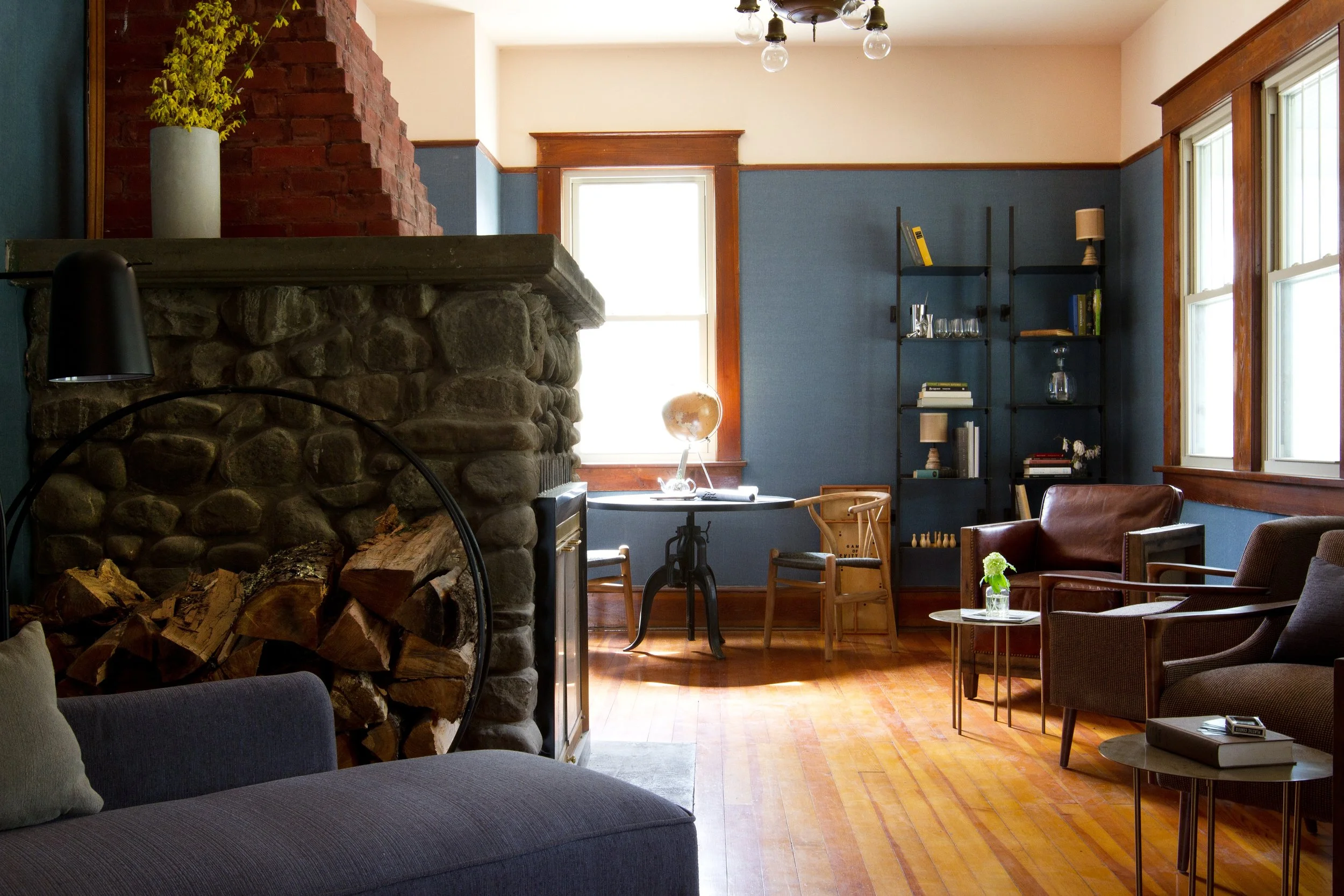Living room with stone fireplace, blue walls, wooden trim, large windows, and a variety of seating and decor including a globe, books, and plants.