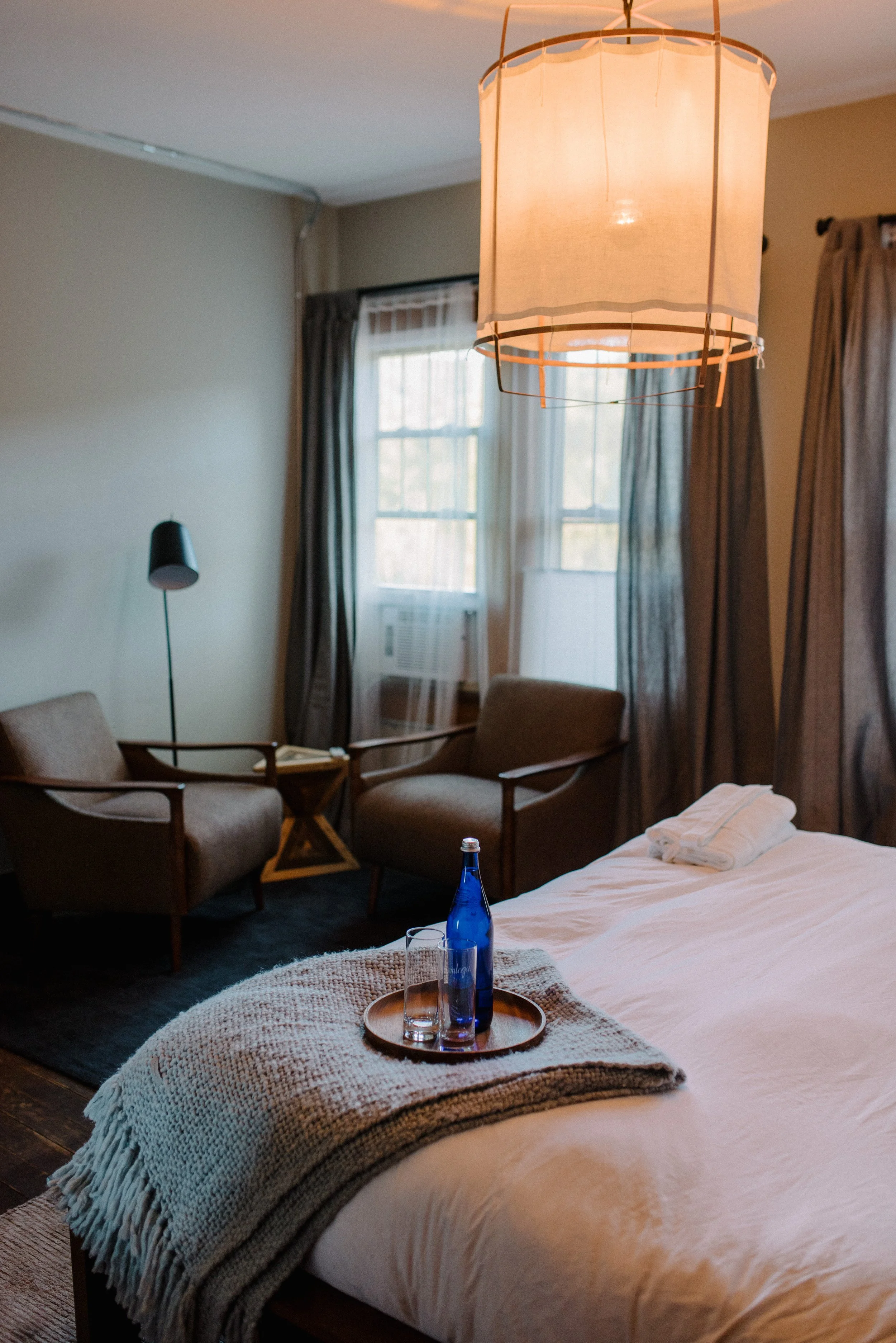 A cozy hotel room with a bed, a tray with water, glasses, and a towel, two armchairs, a side table, and a large hanging lamp, with windows covered by curtains letting in natural light.