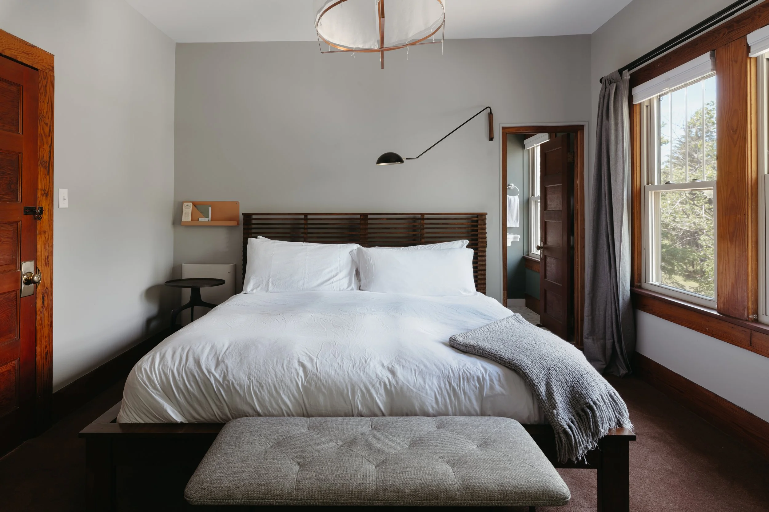 A bedroom with a large bed featuring white bedding and pillows, a gray throw blanket at the foot, wooden window frames, and a small bedside table, with sunlight coming through the windows.