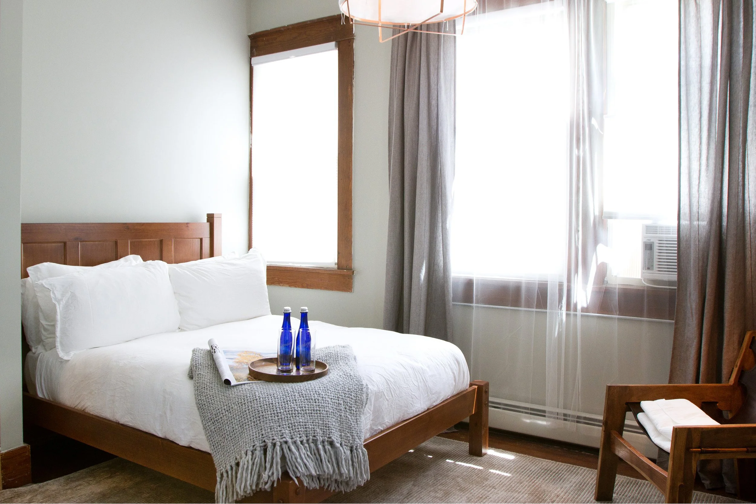 A bedroom with a wooden bed frame, white bedding, and pillows. A tray with two blue bottles, a rolled-up paper, and a gray knitted blanket are on the bed. There are two windows with wooden trim and gray curtains, and an air conditioning unit in the right window. A wooden chair with a towel on it is beside the bed.