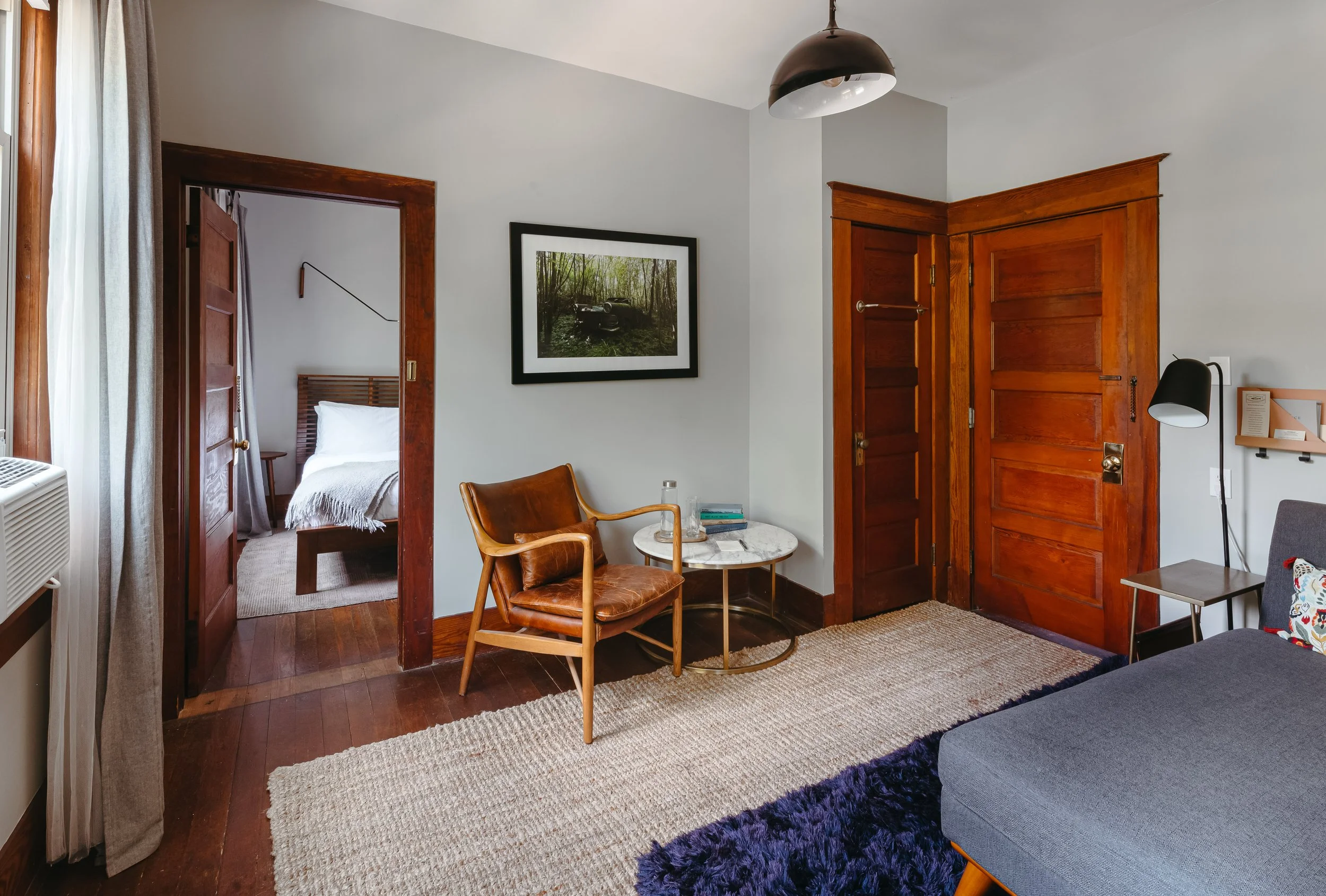 Living room with wooden doors, a gray sofa, a black lamp on a small side table, a chair, and a framed picture on the wall. An open doorway leads to a bedroom with a bed.