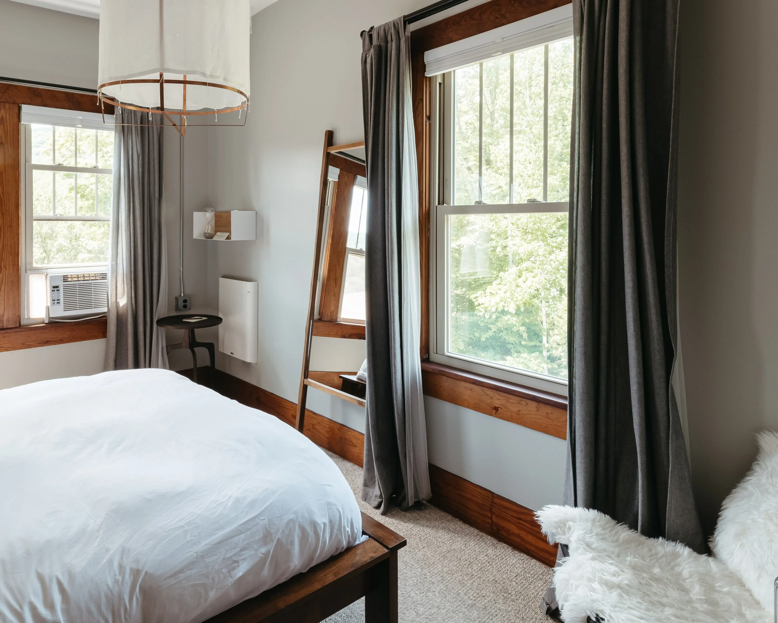 A bedroom with a bed, gray curtains on two windows, a mirror, a small black side table, an armchair with a white fluffy cover, and a wooden frame with a white ceiling light.