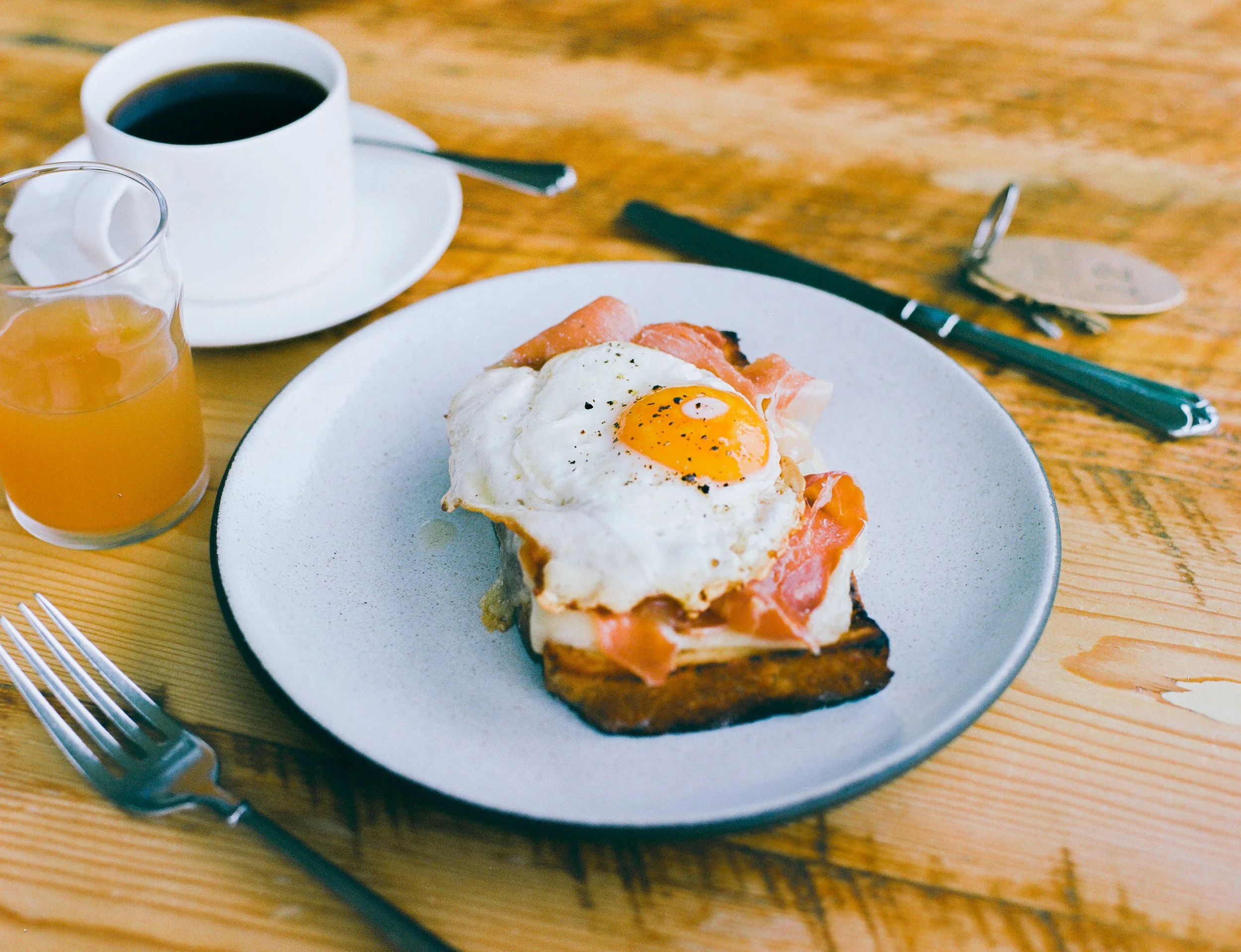 Breakfast plate with fried egg, slices of prosciutto, and toasted bread. Accompanied by a glass of orange juice, a cup of black coffee, and a spoon and fork on a wooden table.