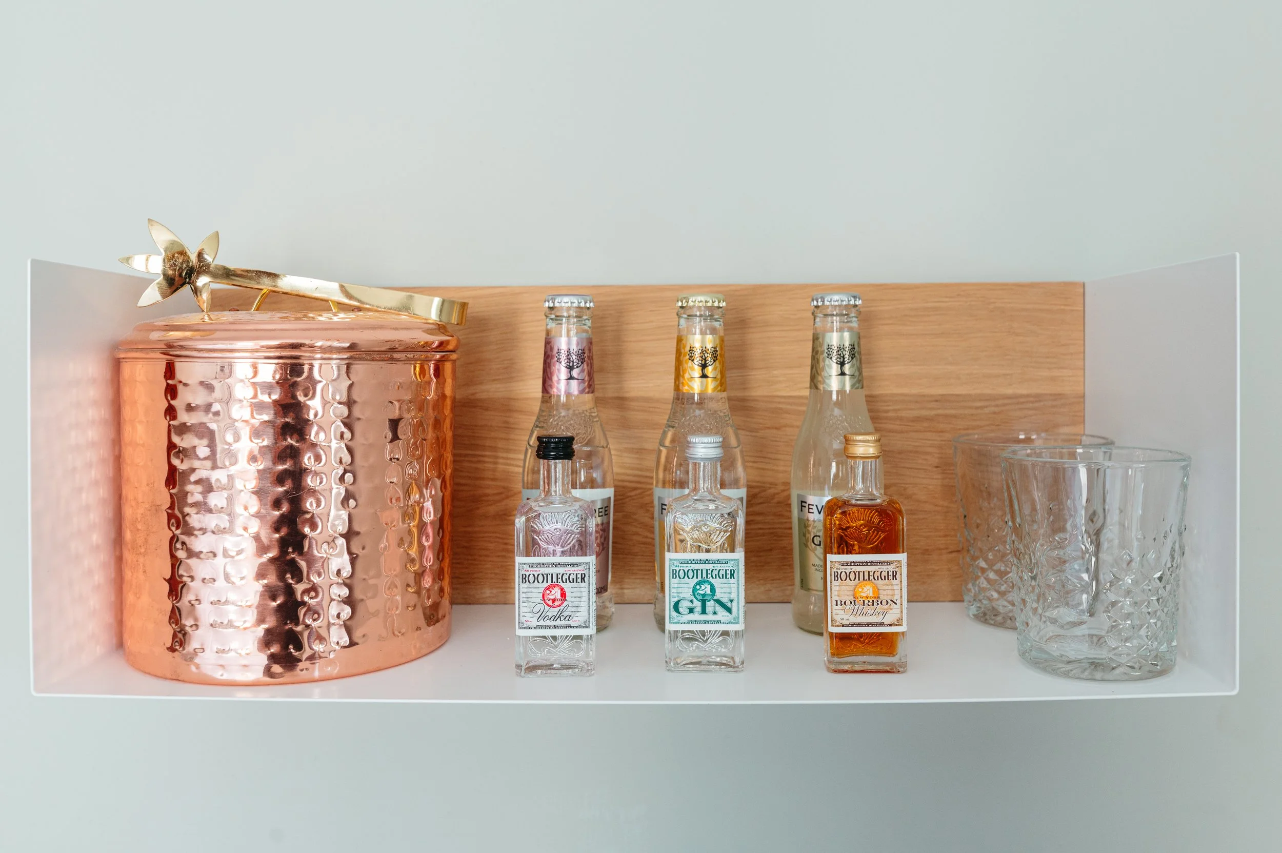 A bar cart shelf displaying mini liquor bottles labeled vodka, gin, and whiskey, a large copper ice bucket with a gold flower bottle opener, and empty crystal glasses with textured patterns.