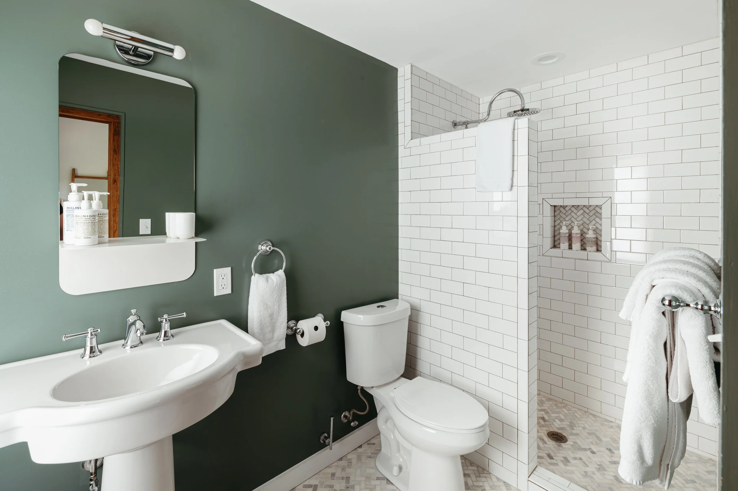 A modern bathroom with a white pedestal sink, a large mirror, green wall, white toilet, and a walk-in shower with white subway tiles and a built-in niche for toiletries.