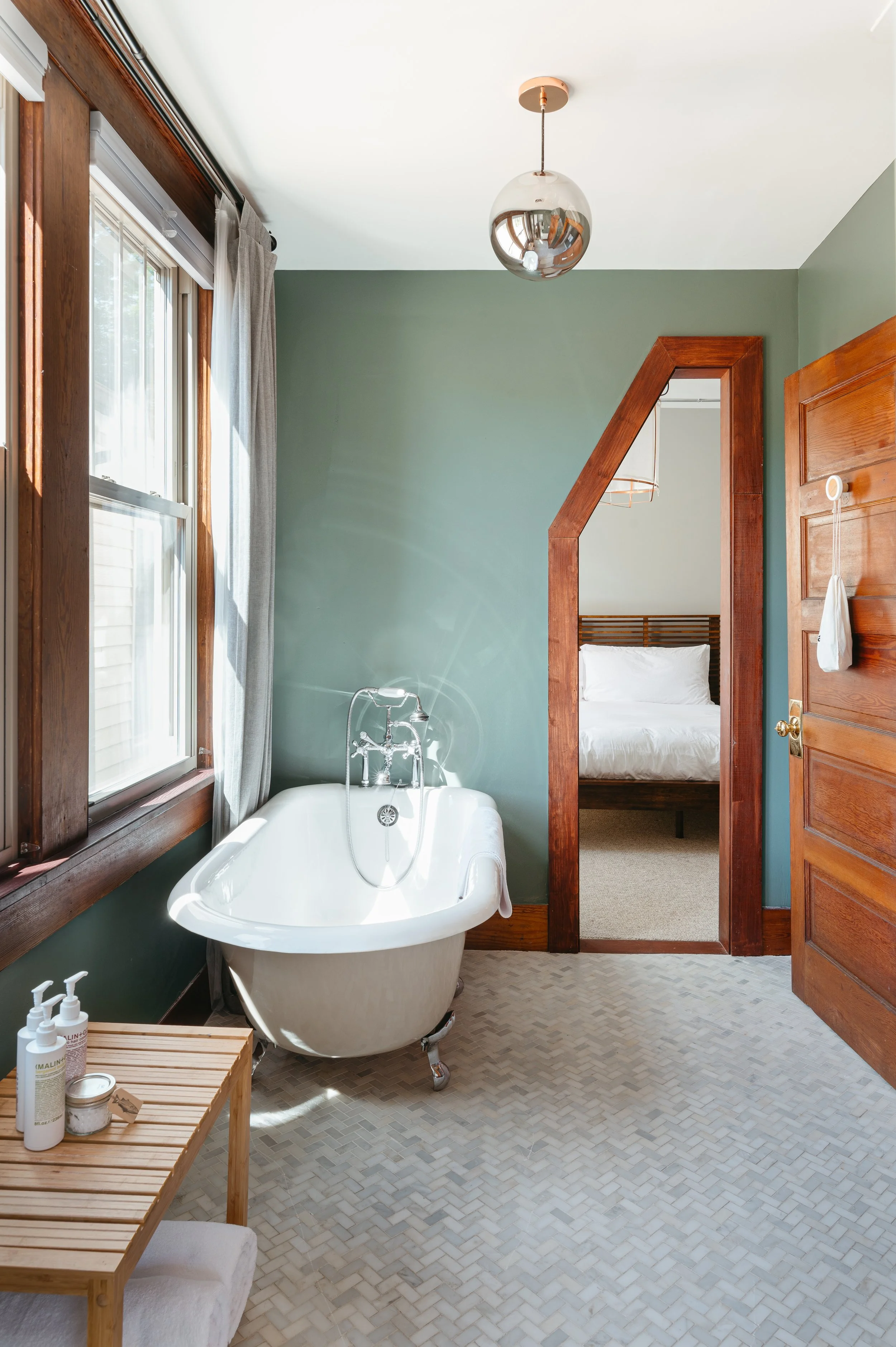 A bathroom with a clawfoot tub near a window, a small wooden table with toiletries, and an open door leading to a bedroom with a bed.