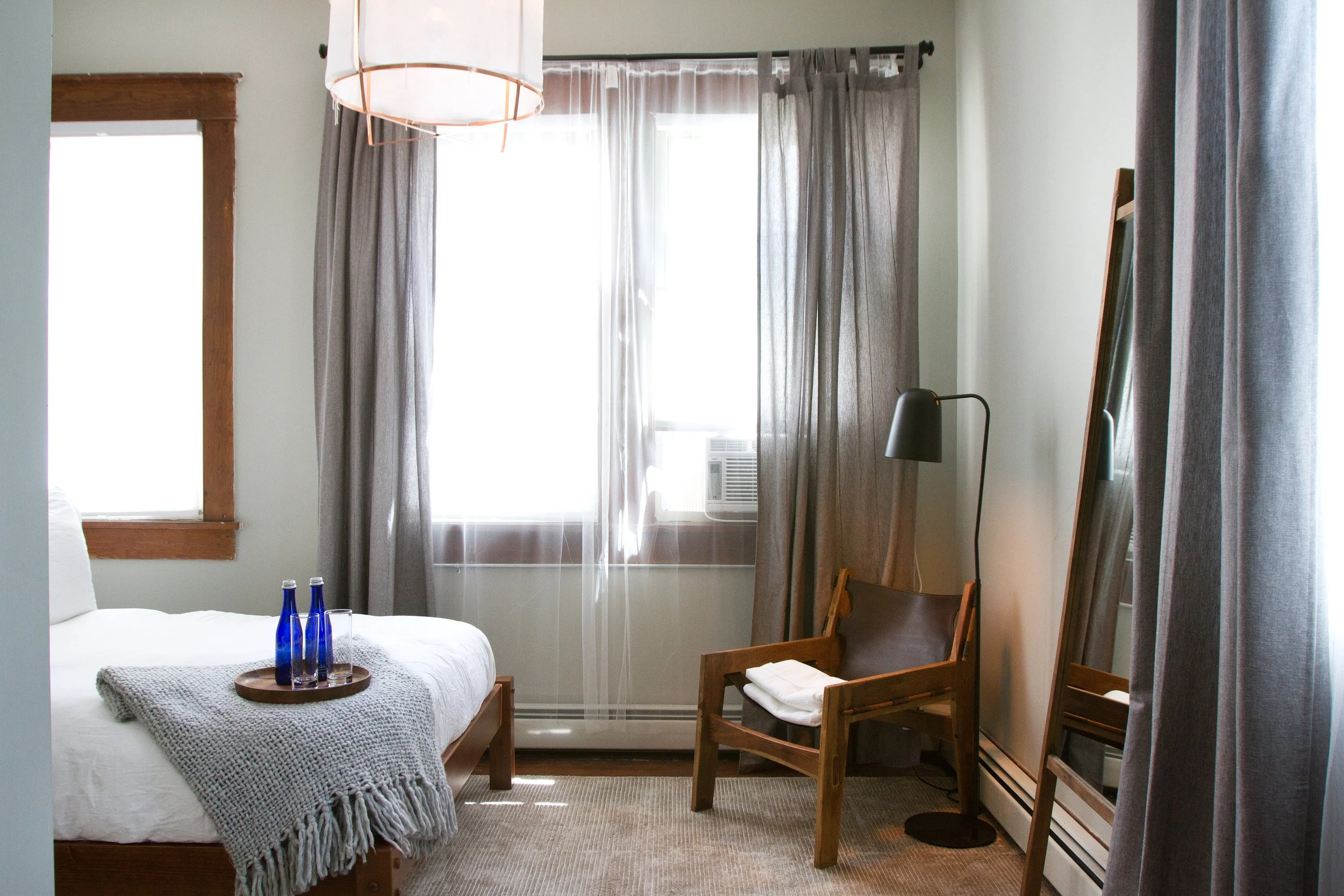 Bedroom with a bed, side table with three blue bottles, wooden chair with a white towel, standing black lamp, and large windows with curtains