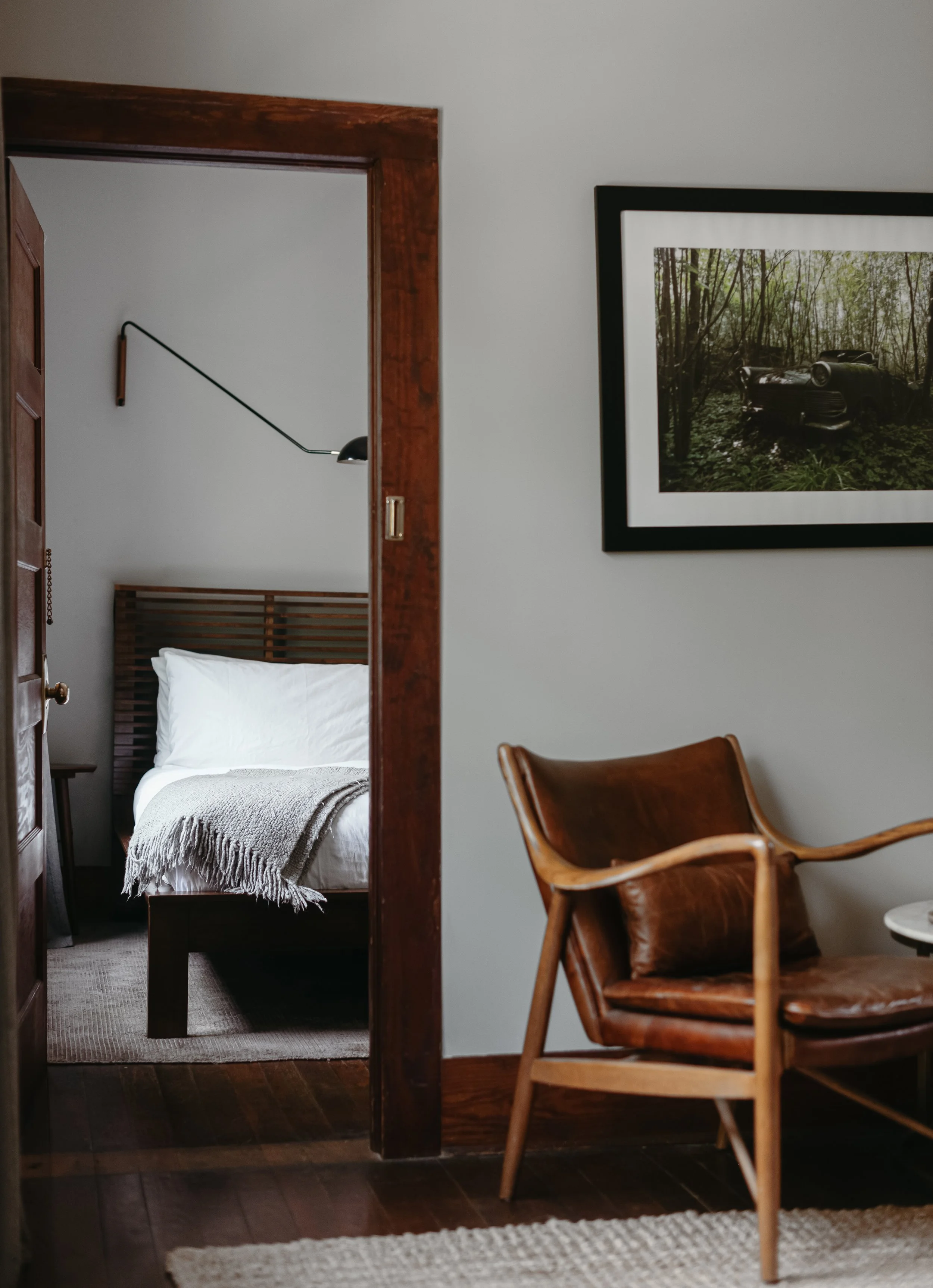 A view through a doorway into a bedroom with a bed, blanket, and wall lamp. In the foreground, there is a brown leather chair and a framed photo of a car in the woods hanging on the wall.