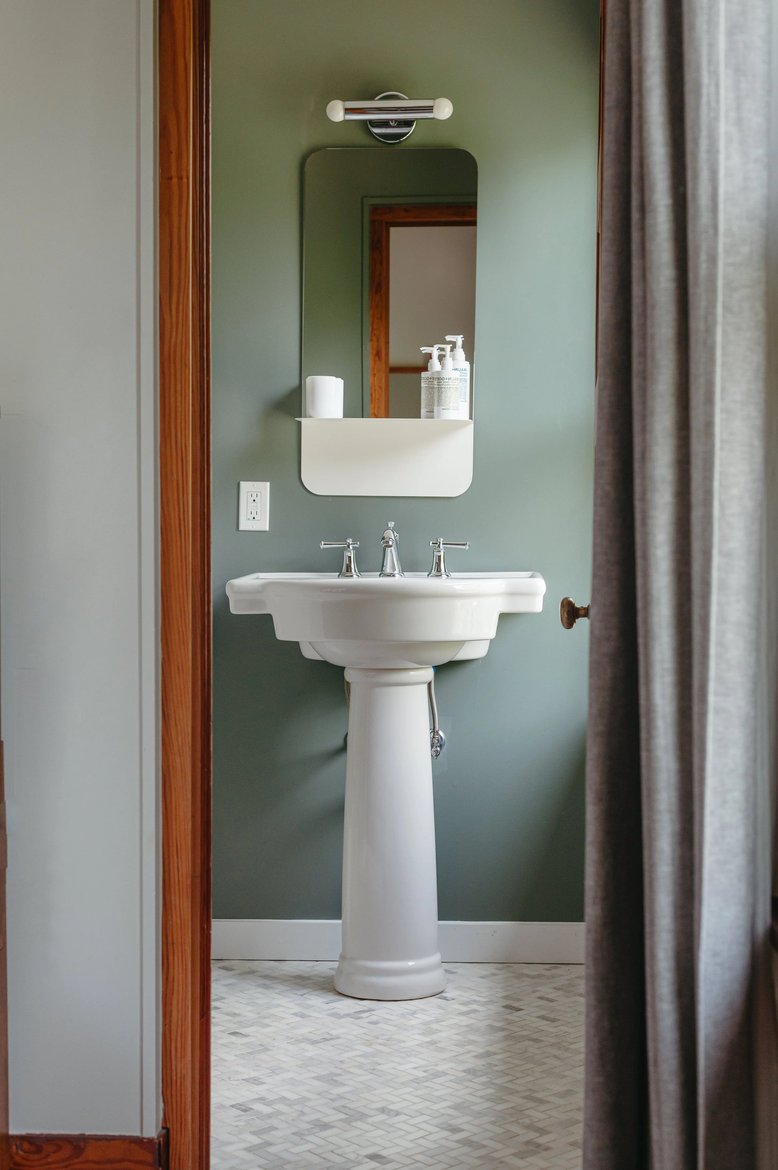 View of a bathroom pedestal sink with a mirror and toiletries, seen through a doorway with curtains on the right.