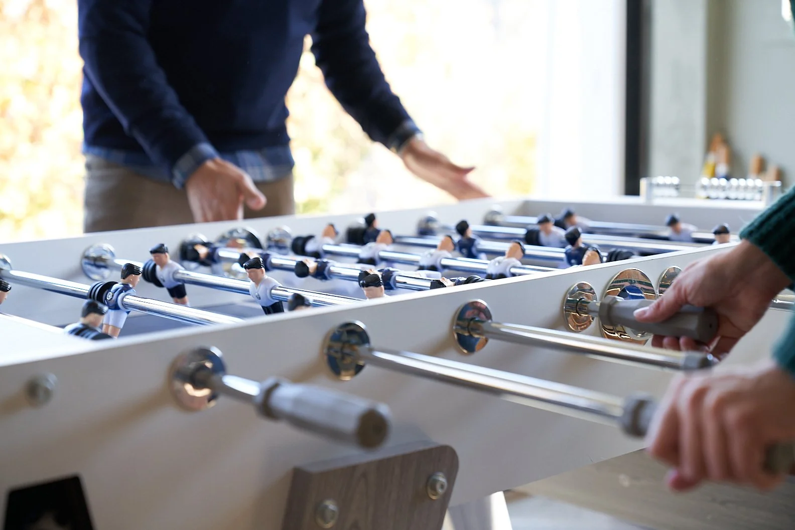 People playing foosball indoors, with a focus on the foosball table and players' hands.