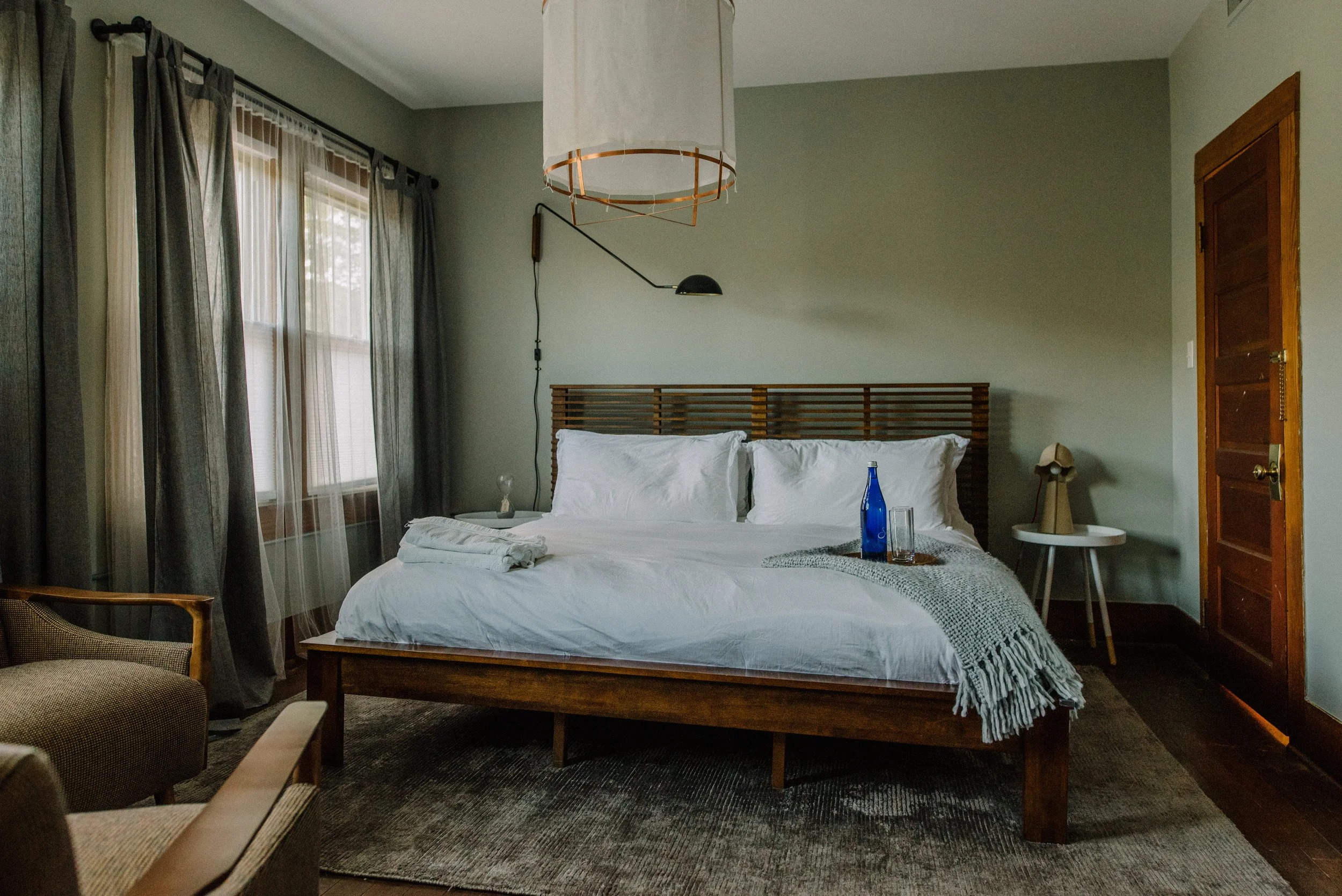 A neatly made bed with white linens and pillows in a bedroom. There is a wooden headboard, a side table with a lamp, and a tray with a blue bottle and glasses on the bed. The room has gray walls, large windows with curtains, and wooden trim.