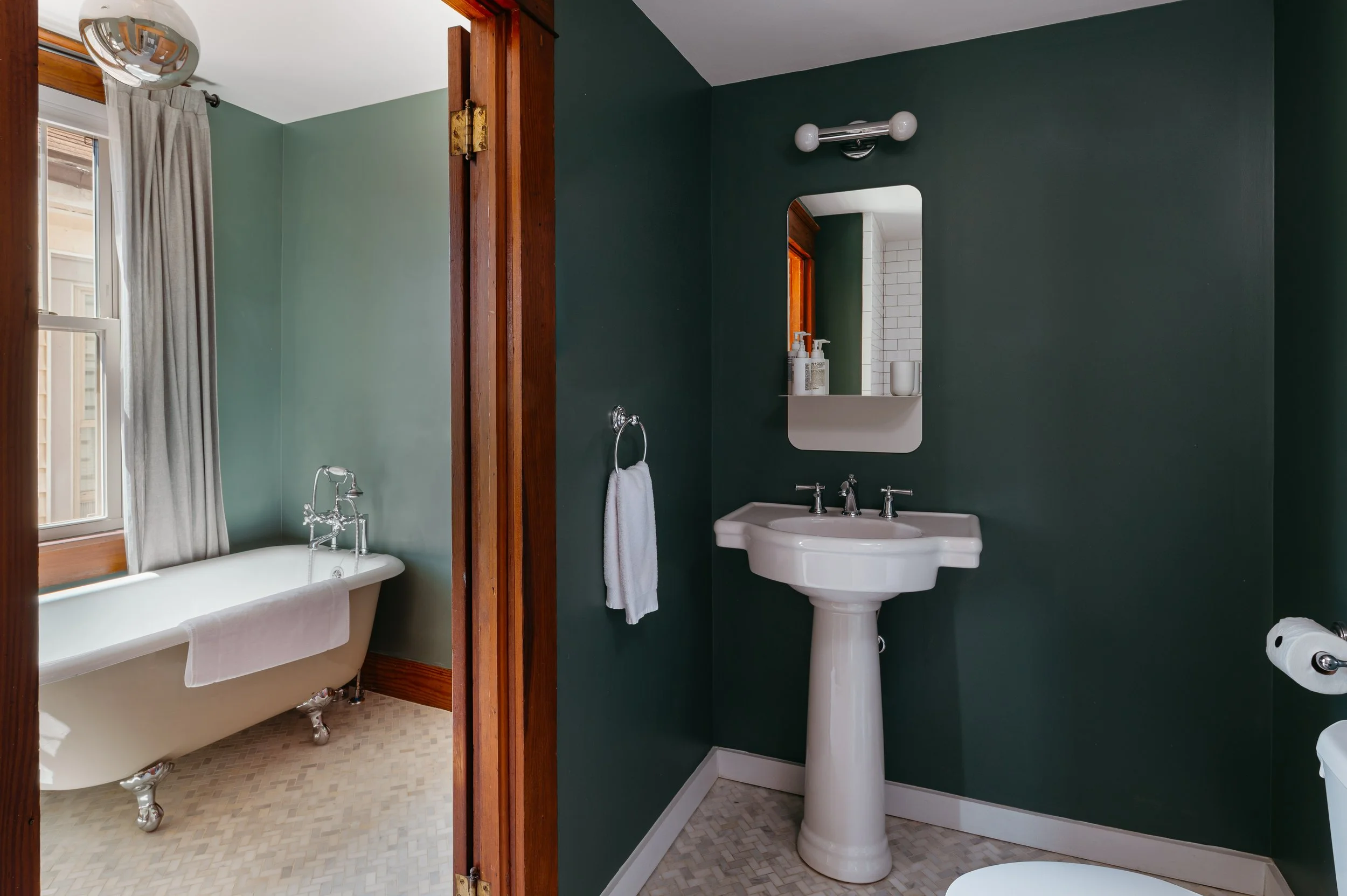 A bathroom with a vintage clawfoot bathtub next to a window with beige curtains on the left side, and a sink with a mirror on the right side. The walls are painted dark green, and the floor has small white tiles. The door frame and window trim are wooden.