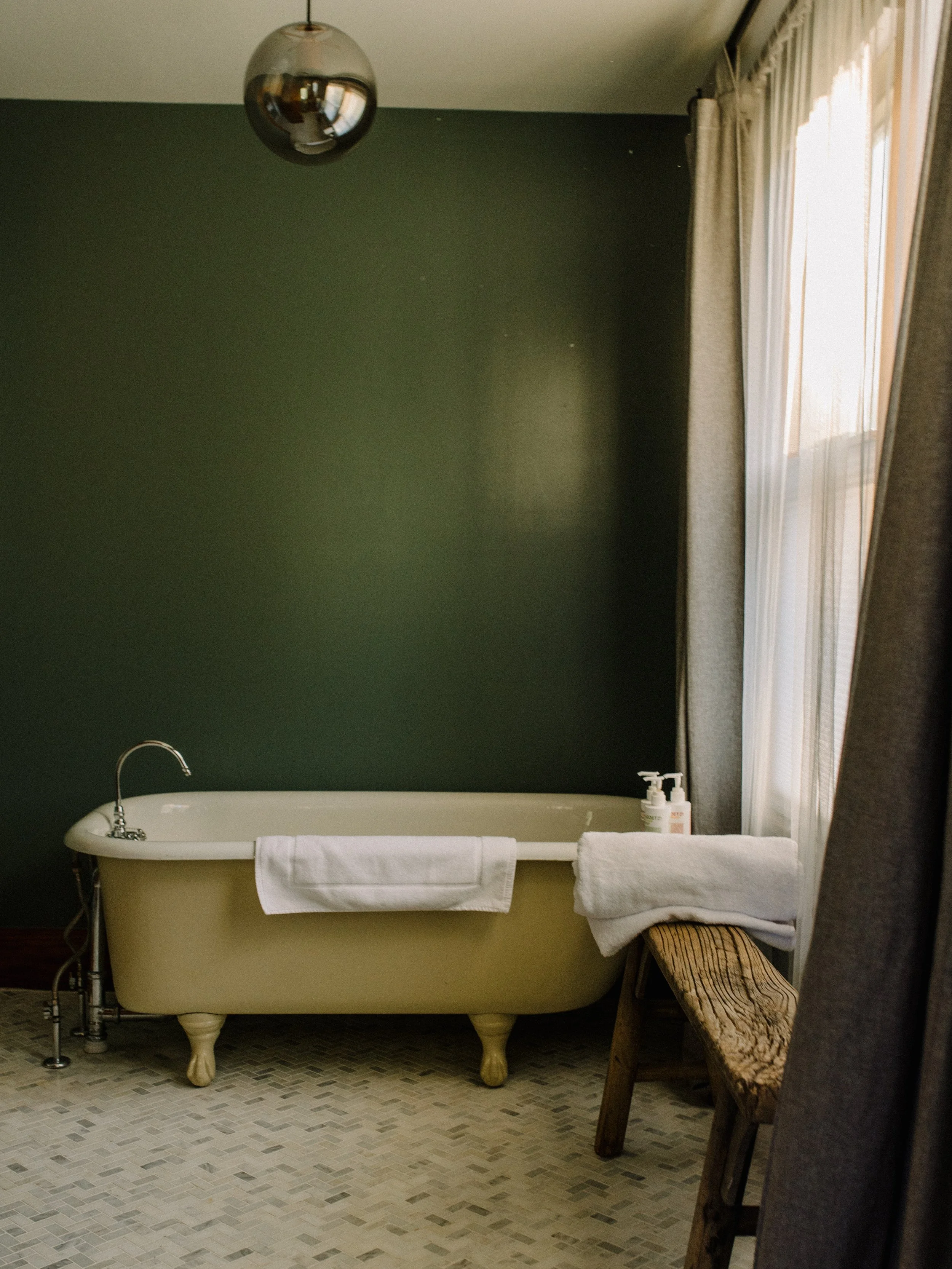 A vintage clawfoot bathtub with white towels hanging on the side, set against a dark green wall in a bathroom, with bottles of soap or lotion on a small wooden bench and a window with curtains letting in natural light.