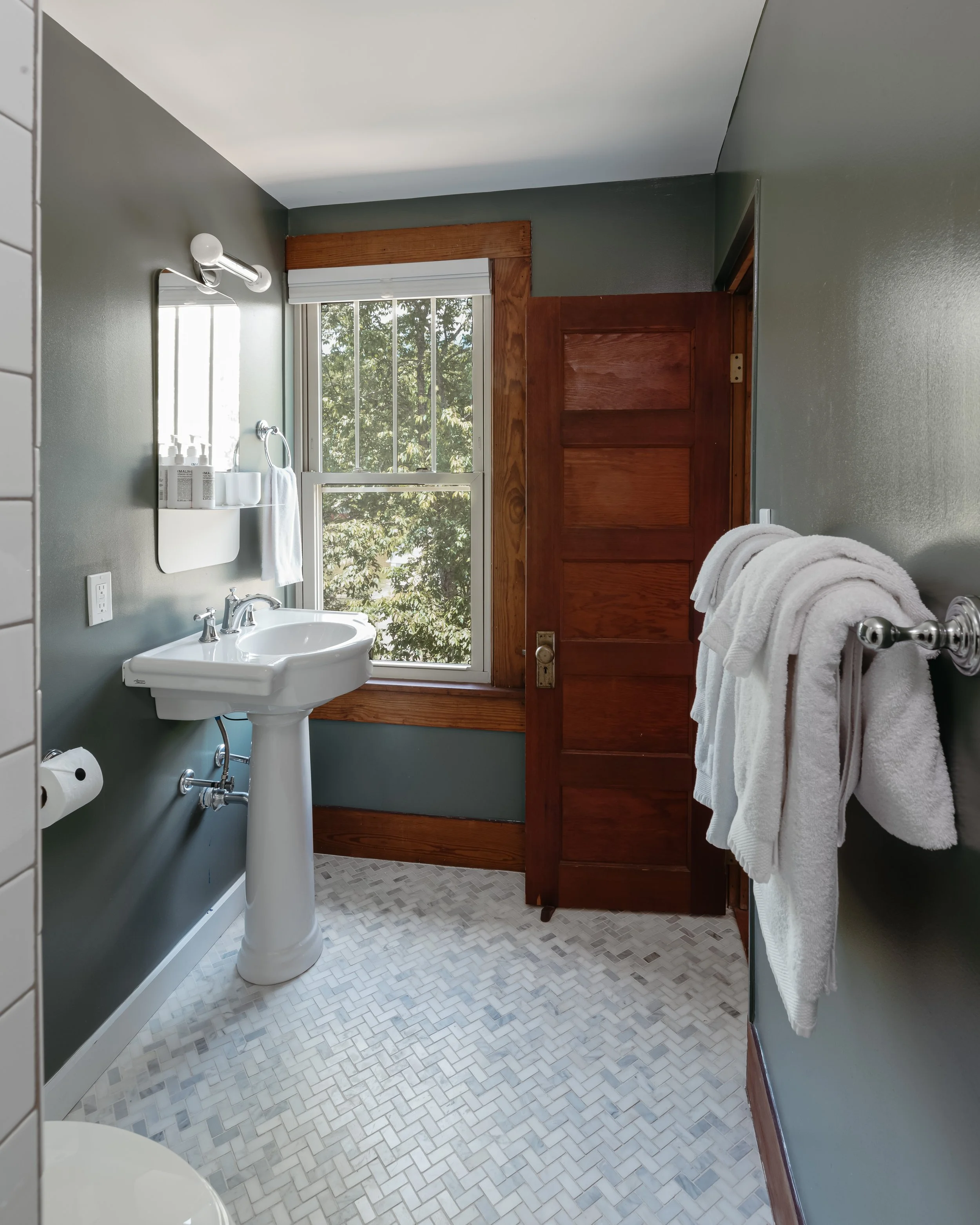 A small bathroom with a white pedestal sink, a towel ring with a white towel, a mirror above the sink, a window with trees outside, a wooden door, a towel rack with white towels, and tile flooring.