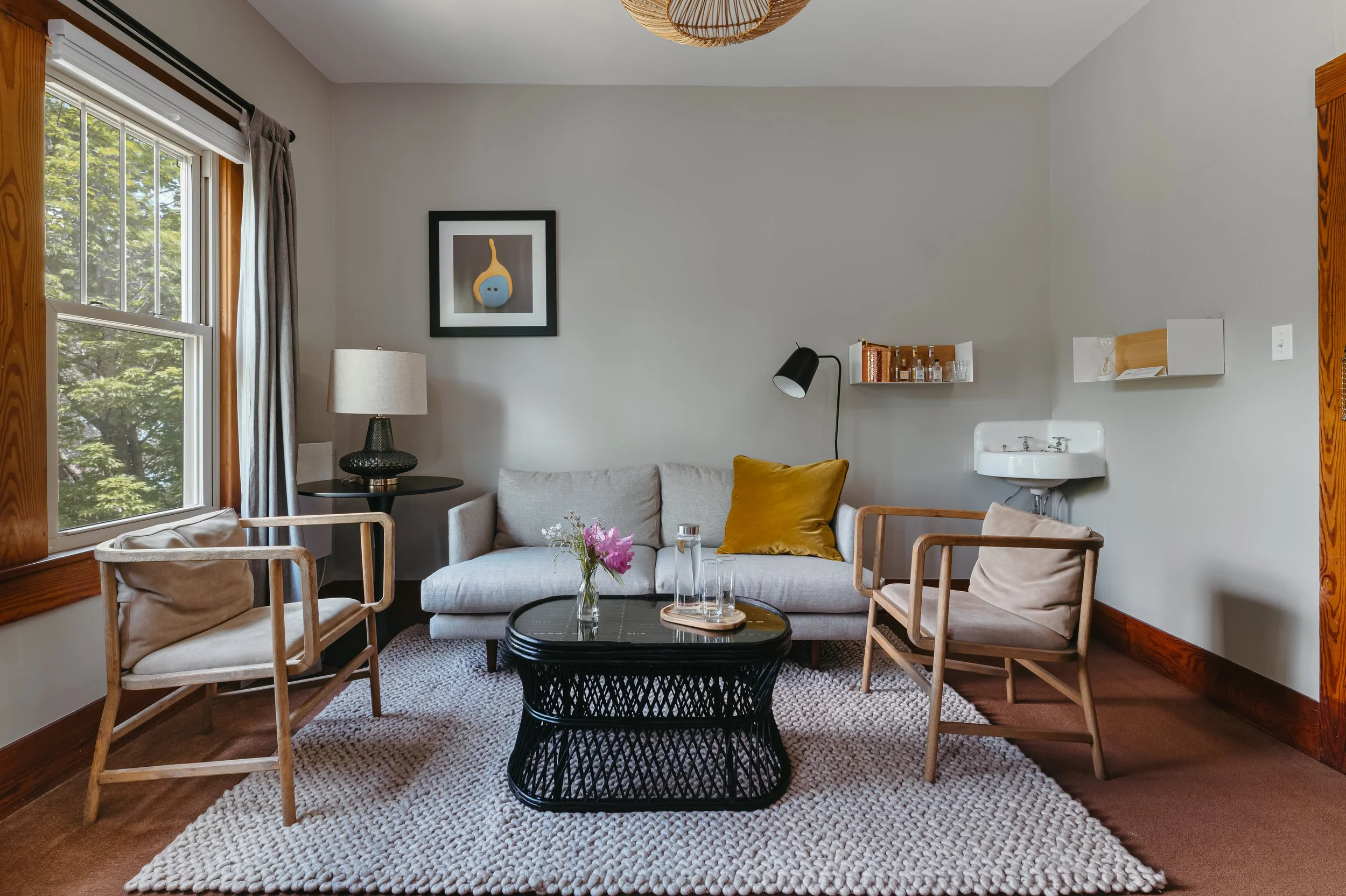 Living room with gray sofa, two wooden armchairs with beige cushions, black coffee table with flowers and glasses, window with gray curtains, wall art, bedside lamp, small sink on the wall, and a textured beige rug.