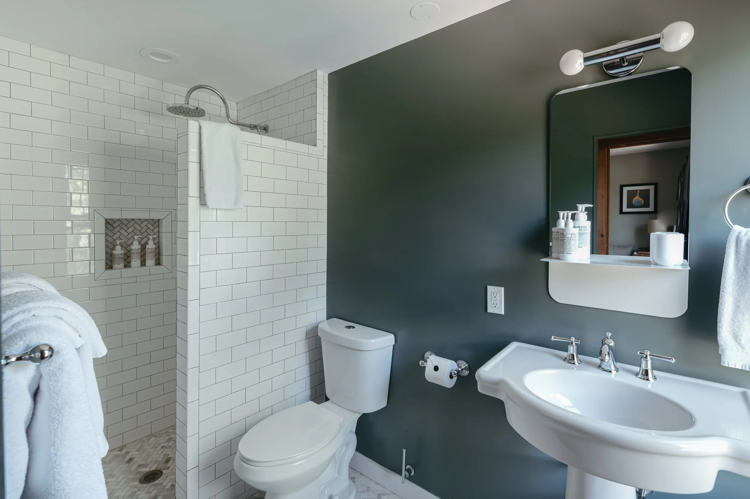Bathroom with shower, toilet, and sink, decorated with white tiles and gray wall, including bottles of soap on the shelf and towels.