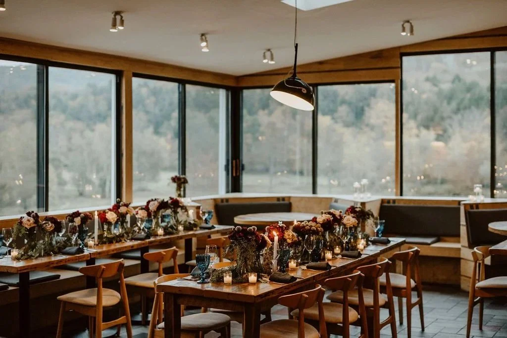 Indoor dining area with a wooden table set with flowers, candles, and tableware. Large windows reveal an outdoor view of trees and hills.