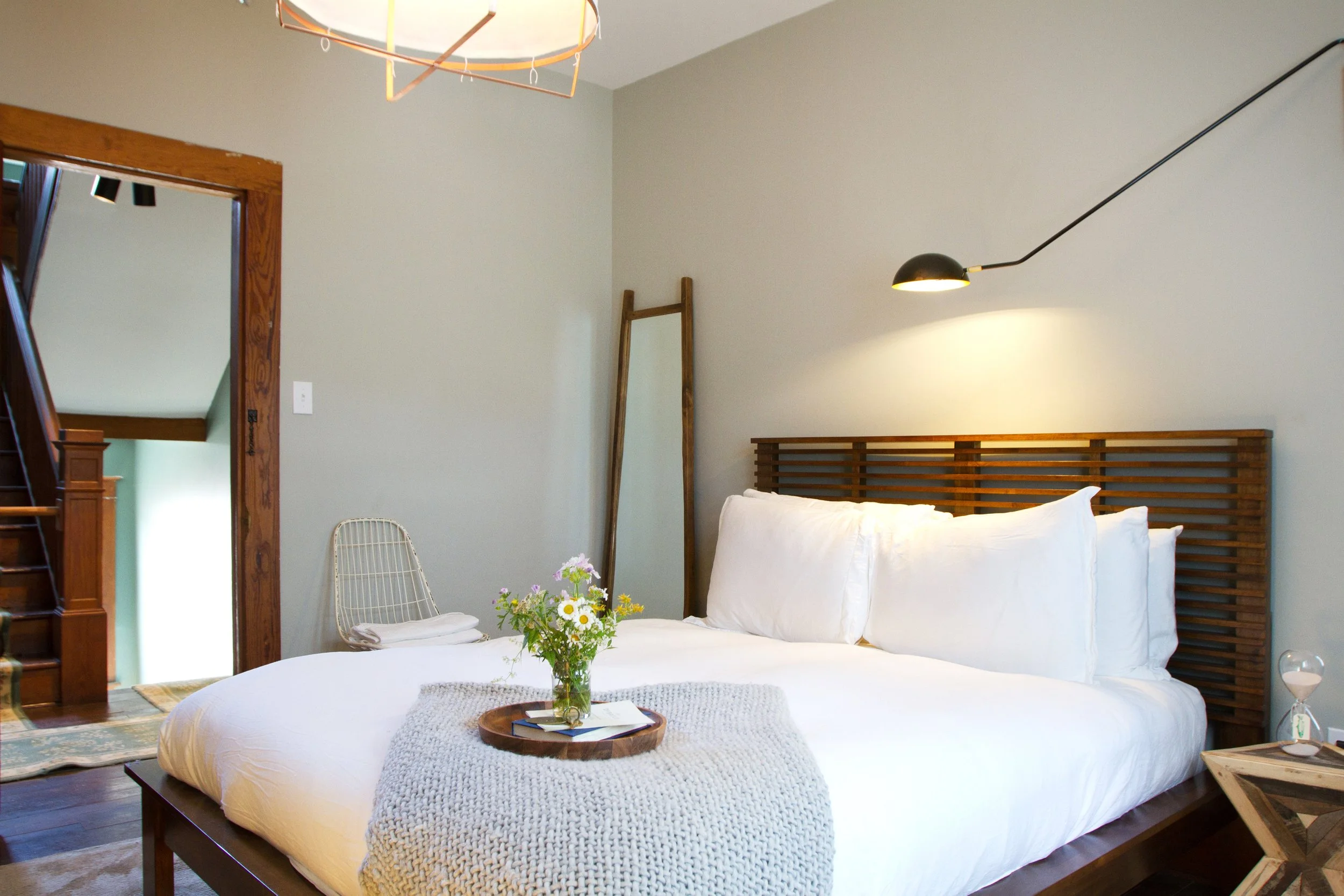 A cozy bedroom with a large bed featuring white bedding and pillows, a wooden headboard, a side table with an hourglass, a flower vase on a knitted blanket, a chair with folded towels, a full-length mirror, and a wall-mounted black reading lamp, with a doorway leading to a staircase.