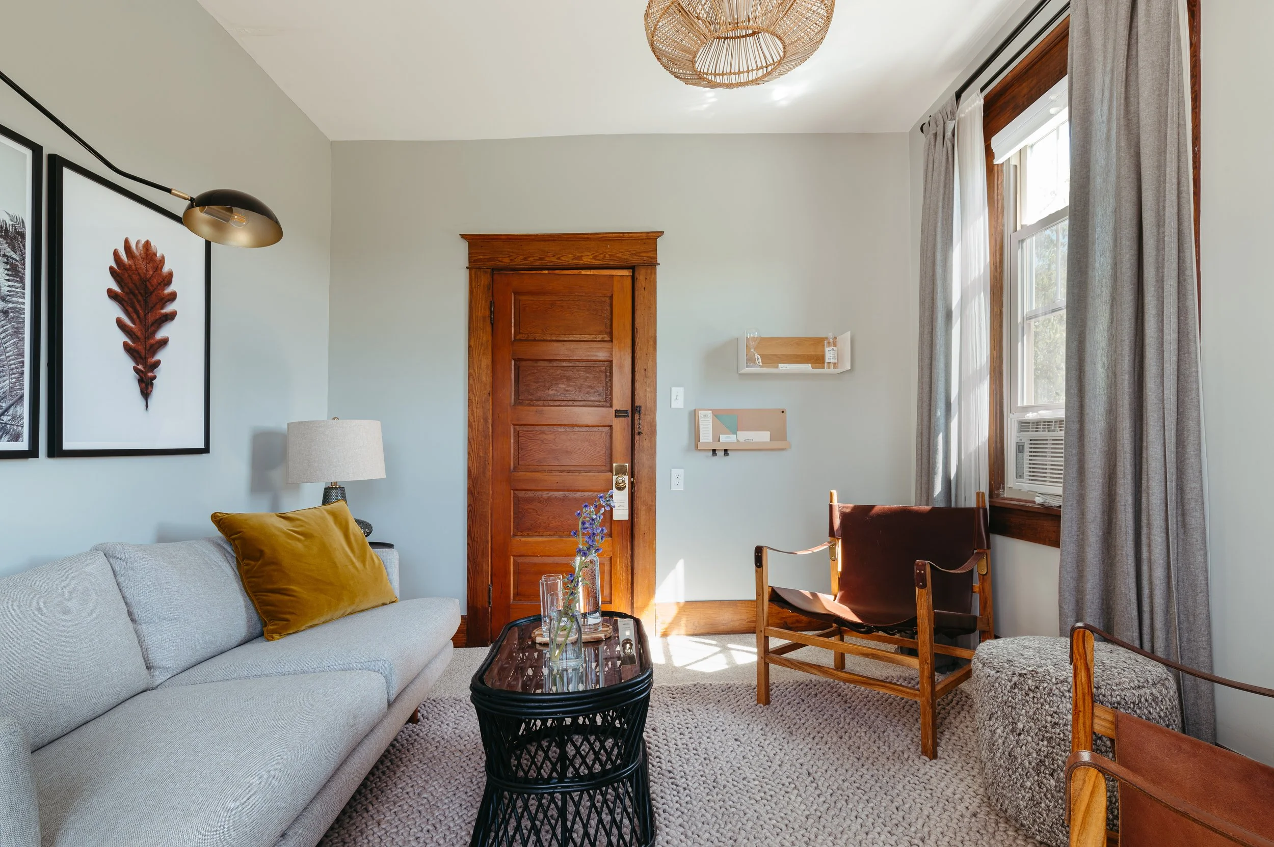 Living room with a light gray sofa, two artwork frames, a black side table with a glass top, a gold pillow, a table lamp, a wooden door, a wall-mounted shelf, flowers on the table, a window with gray curtains, an air conditioning unit, a leather and wood chair, a chunky ottoman, and a patterned carpet.