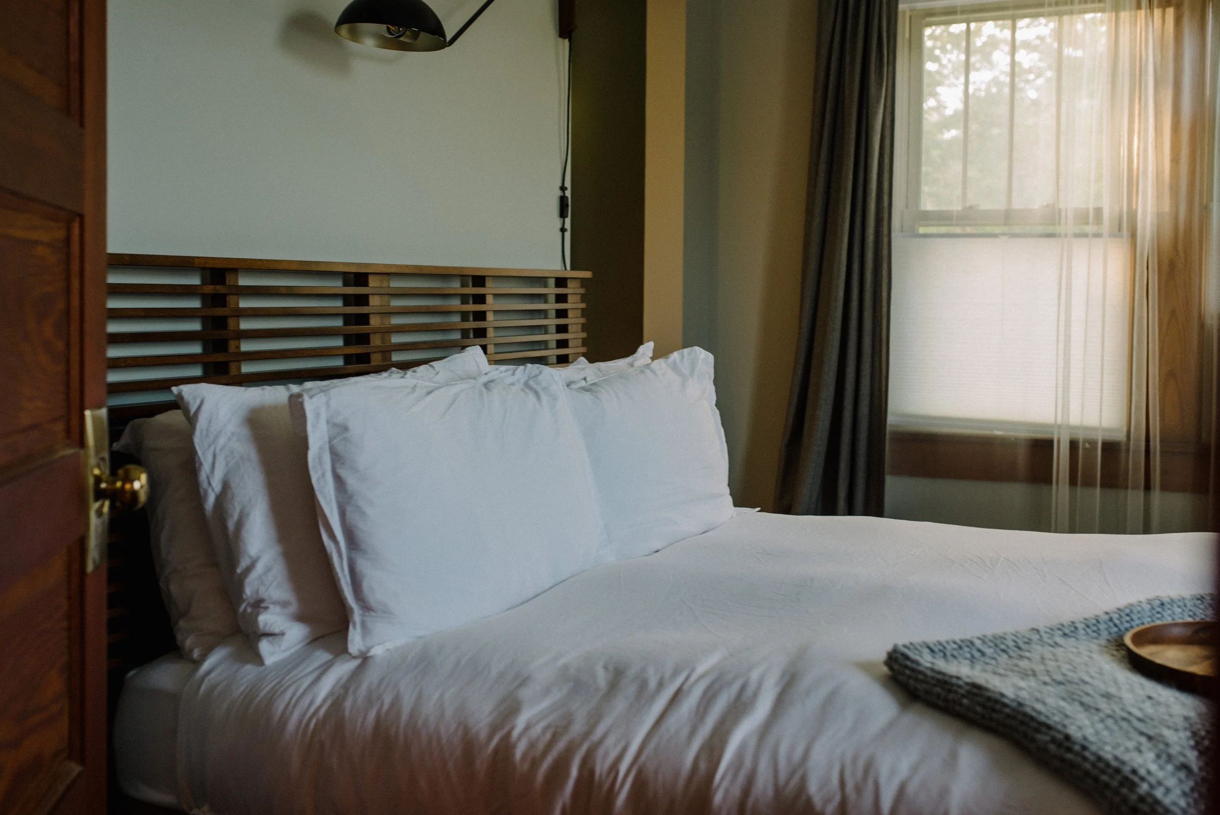 A neatly made bed with white sheets and pillows in a bedroom with natural light coming through a window with dark curtains.