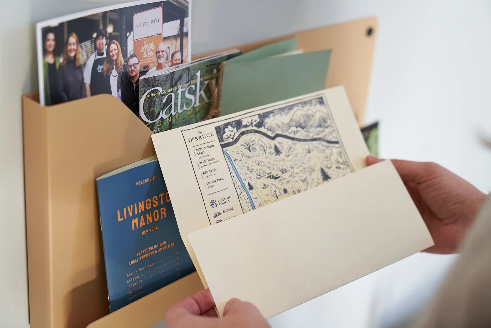 A person's hands hold a beige wall-mounted brochure holder filled with various brochures and magazines, including one titled 'Livingston Manor' and others with images and maps.