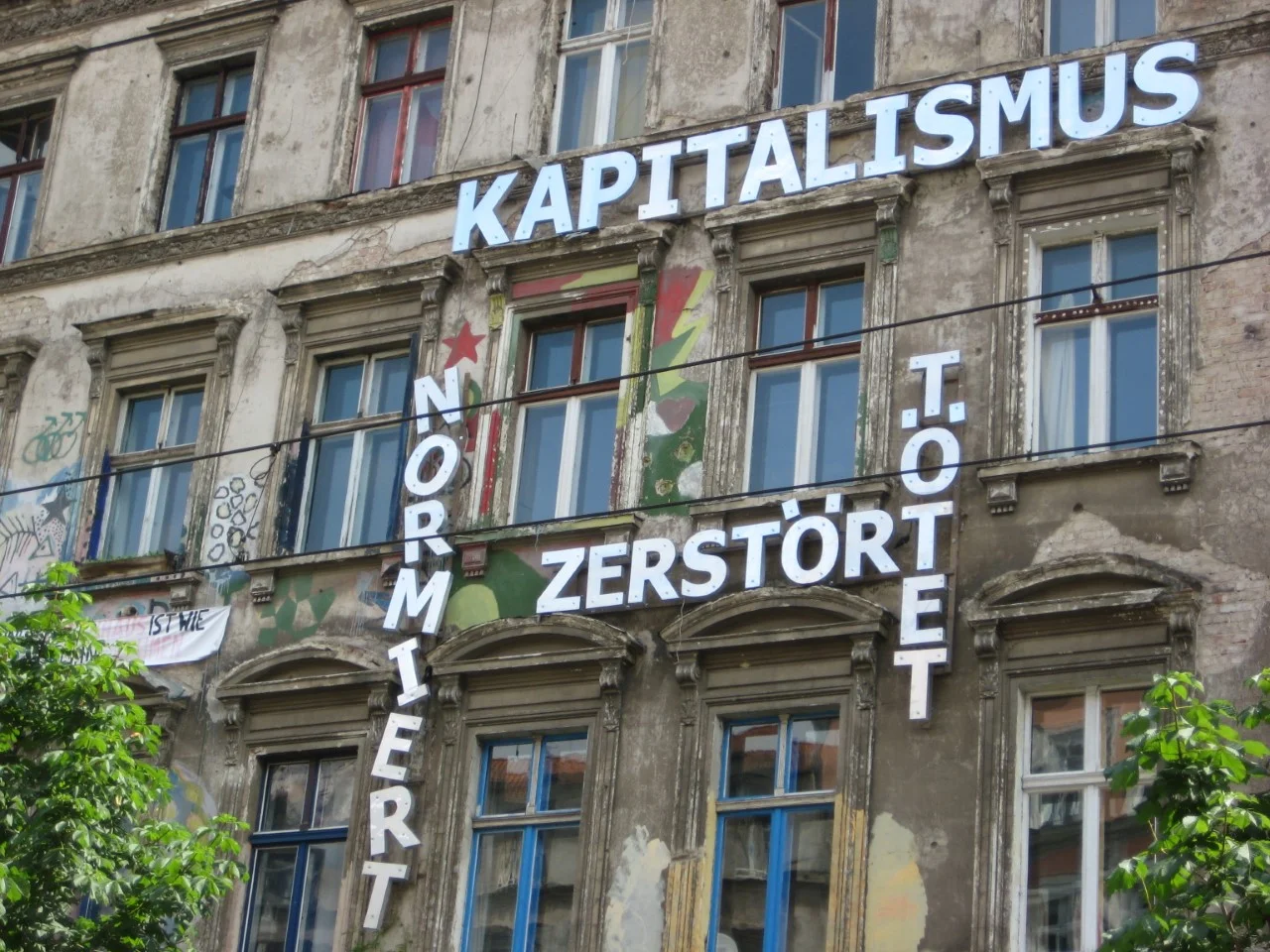 A message on the facade of Kastanienallee 86 reads "Capitalism normalizes, destroys, kills". The building contains two Hausprojekte, or “living projects” that proliferated the new city center of reunified Berlin: the Tuntenhaus, or “Queer House”, takes up the back wing of the building, and the KA 86 Hausprojekt faces the street. 2007. Courtesy of Daniela Sandler.
