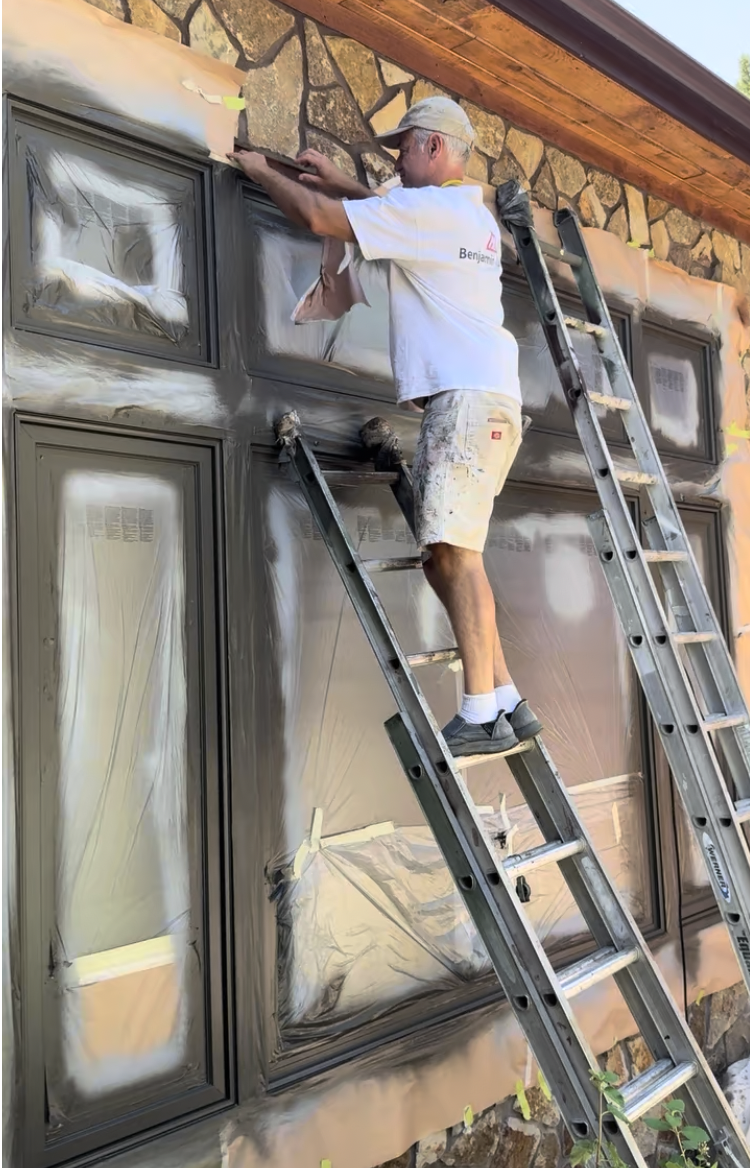 A man standing on a ladder painting the exterior of a house, focusing on the window frames, with masking tape and plastic covering the surrounding areas.