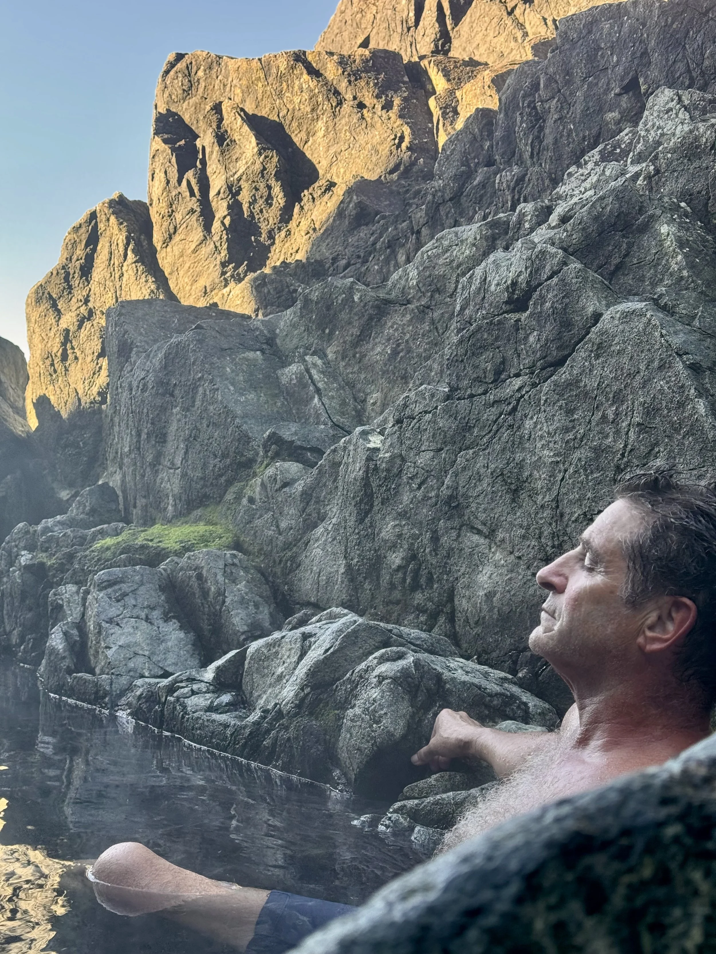 A man relaxing in a hot spring or natural pool surrounded by large rocks and cliffs, with the sky visible in the background.