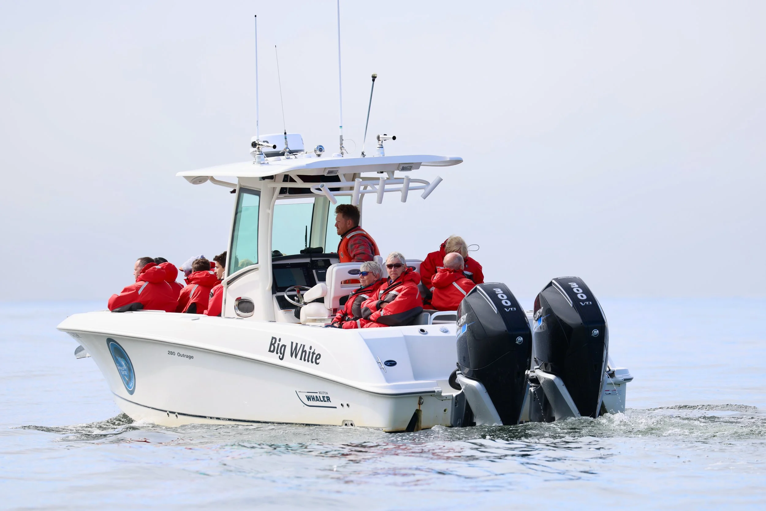 A group of people in red jackets on a white boat named 'Big White' with two outboard motors, sailing on calm water under a cloudy sky.